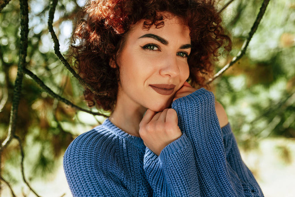 Outdoor close-up portrait of gorgeous young woman with curly hair, smiling and looking to the camera, posing against nature background, has positive expression, wearing blue knitted sweater. - 写真・画像