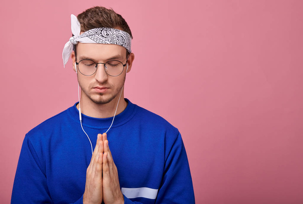 Confident guy with glasses in a white bandana and a blue sweater to listening to music with earphones in his ears. Boy's arms folded, as if praying. Pink wall back, copy space. Focus on hands. - Foto, immagini