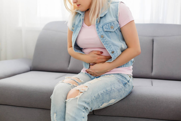 期間を持つアジアの女の子はソファの上に座っていると彼女の胃の上に痛みを感じる。女性の痛みや月経症候群と呼ばれています。彼女は胃の上に保持し、痙攣を通してカール. - 写真・画像