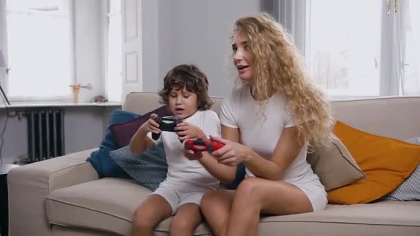 Pretty cheerful blond woman with curly hair playing video game with her attractive little son using joysticks,sitting on the cozy couch in the room - Záběry, video