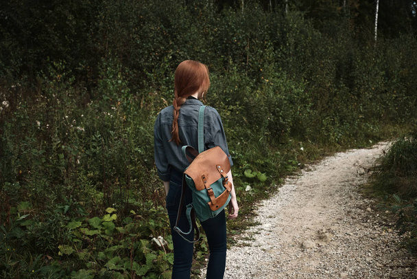 A red-haired girl is walking in the forest along the road with a backpack, a frame from the back from the side. - Photo, image