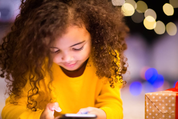 Aranyos kis afrikai lány játszik mobiltelefonnal karácsony este - Fotó, kép