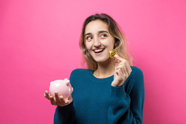 Mädchen legt in das Sparschwein physische bitcoin.young Mädchen über rosa Hintergrund hält Sparschwein und jubelt gestikulierend. das Konzept der Zuverlässigkeit von Geldanlagen und Versicherungen. - Foto, Bild