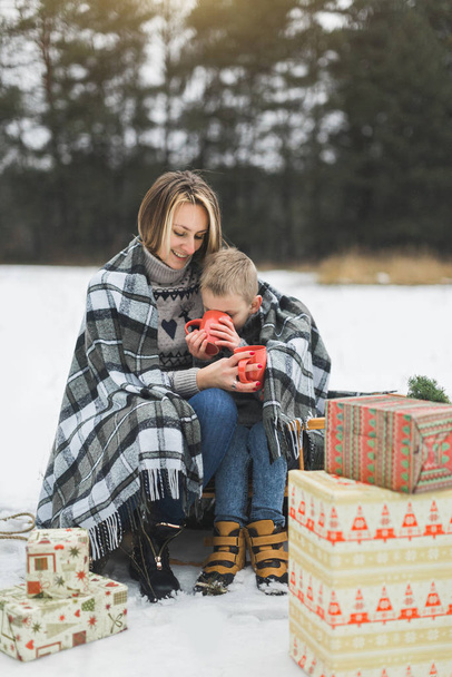 Little boy and mother sliding in the winter forest. Mom and son sitting on the sledge, covering by the checkered plaid and drinking hot drinks. Decorated present gifts around them - Фото, изображение