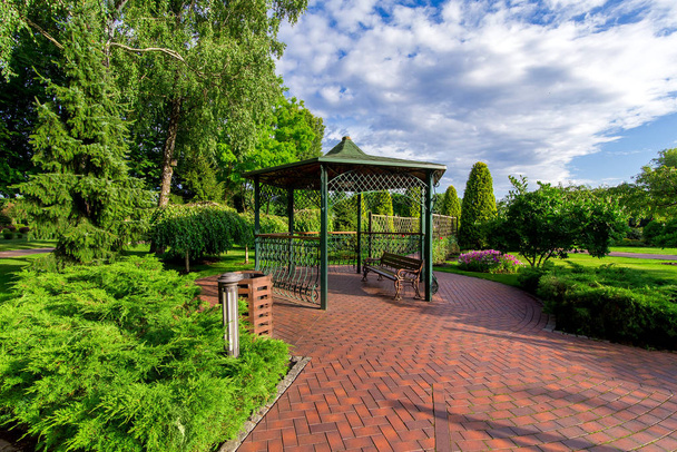 an iron gazebo with a bench and paving stone tiles in the garden with a lawn and evergreen thuja on a sunny day with a cloudy sky, nobody. - Fotografie, Obrázek