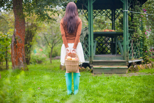 Rear view of young woman in the garden of bright rubber boots in hands of straw basket with apples - Photo, Image