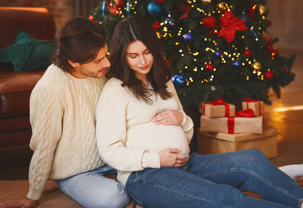 Беременная женщина, сидящая у рождественской елки со своим мужем
 - Фото, изображение