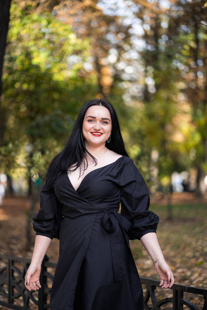 Autumn fashion outdoor. The girl with red lips in fashionable stylish black dress, autumnal lifestyle on the background of blurry yellow-green trees in the park. Vertical - Foto, Imagen