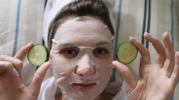 A young woman in a cleansing mask and a towel looks at the camera and holds round cucumbers in her hands. Wellness, beauty product and skincare - Filmagem, Vídeo