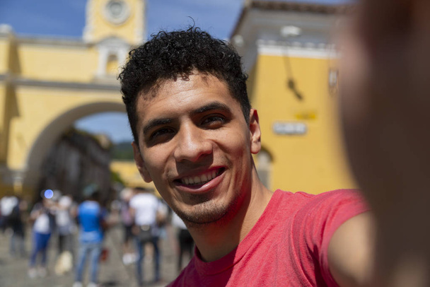 Young Hispanic man taking a self in the city of Antigua Guatemala with the Santa Catalina arch behind - Photo, Image