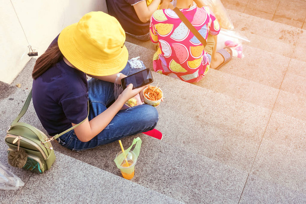 Žena se žlutým kloboukem sedí hrát na smartphone, zatímco druhá ruka drží šálek masa na schodech ve městě - Fotografie, Obrázek