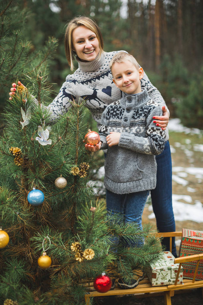 Family, young mother and little son, wearing knitted sweaters, decorating fir tree in the forest or park. Colorful decorations, wooden sledge, Christmas presents - Фото, изображение