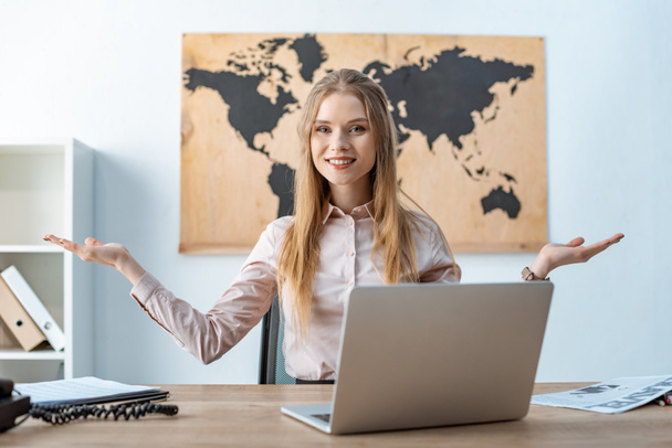 smiling travel agent showing welcome gesture while sitting near laptop - Photo, Image