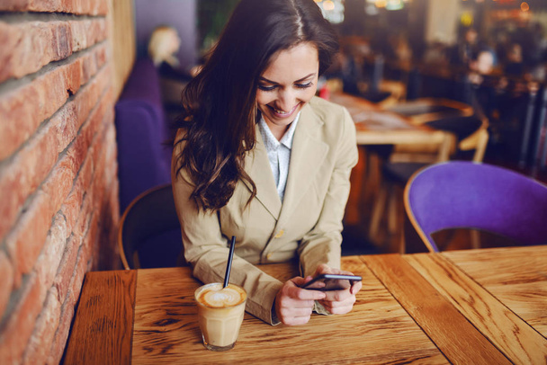 Portret van een prachtige Kaukasische brunette gekleed slim casual zitten in cafe en met behulp van de smartphone voor het verzenden van bericht. Op houten tafel is verse heerlijke koffie. - Foto, afbeelding
