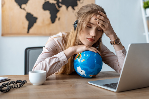 tired travel agent leaning on globe while sitting at workplace with closed eyes - Photo, Image