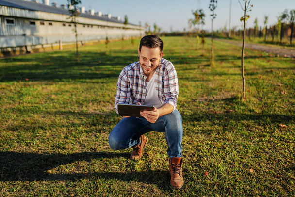 Веселый кавказский фермер в клетчатой рубашке, приседающий на улице и пользующийся планшетом. На заднем плане амбар и сад
. - Фото, изображение