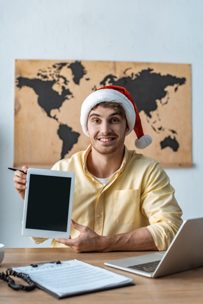 fröhliches Reisebüro mit Weihnachtsmütze, das in die Kamera schaut, während es ein digitales Tablet mit leerem Bildschirm hält - Foto, Bild