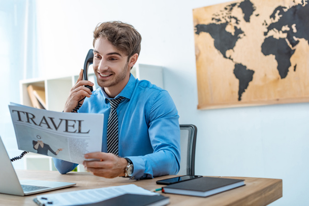 smiling travel agent talking on phone while holding travel newspaper - Photo, image