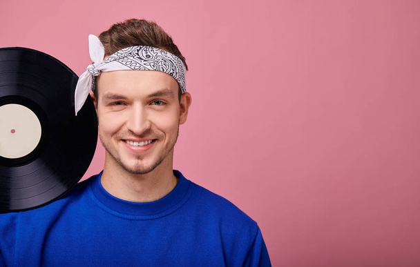 Close-up. A cool stylish guy in a bandana in a dark blue jacket stands on a pink background with a black vinyl record in his hand. Youth, style, confidence, joy, playful mood. Happy. Retro. - Zdjęcie, obraz
