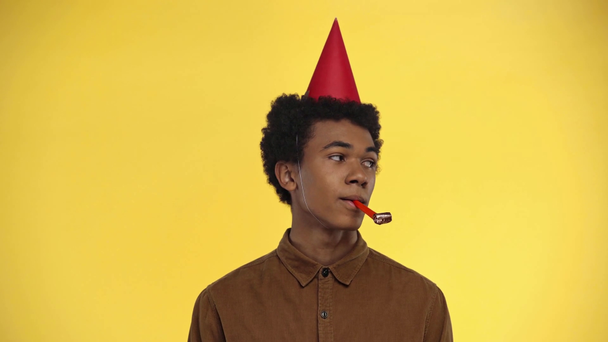 african american teenager blowing in party horn isolated on yellow - Imágenes, Vídeo
