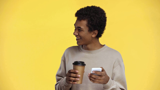 africký americký teenager pití a použití smartphone izolované na žluté - Záběry, video