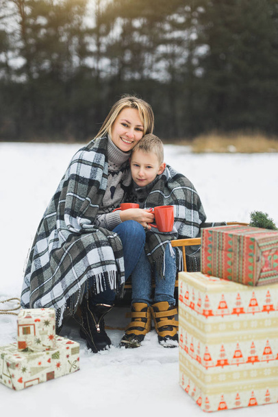 Little boy and mother sliding in the winter forest. Mom and son sitting on the sledge, covering by the checkered plaid and drinking hot drinks. Decorated present gifts around them - 写真・画像