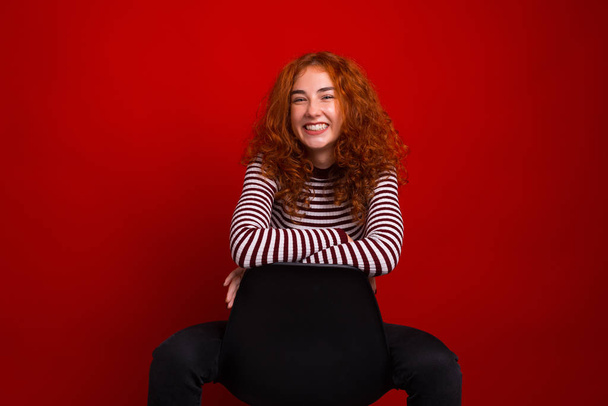 Boldog vörös fiatal nő, ül a széken, és mosolyog a kamerába - Fotó, kép