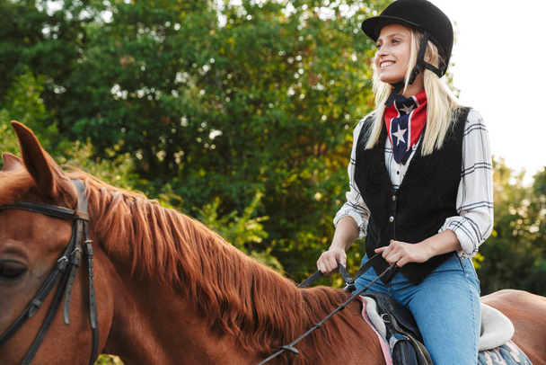 Image of joyful woman wearing hat riding horse at yard in countr - Zdjęcie, obraz