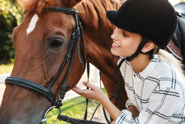 Image of excited woman wearing hat standing by horse at yard in  - Фото, зображення