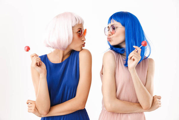 Portrait of two nice funny women wearing wigs and eyeglasses holding lollipops while making kiss face - Fotografie, Obrázek