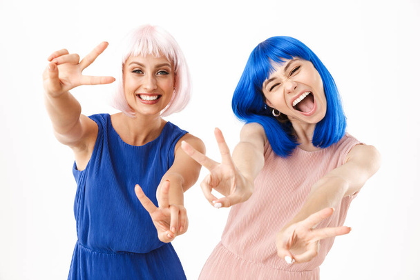 Portrait of two excited women wearing blue and pink wigs smiling while gesturing peace sign - Φωτογραφία, εικόνα