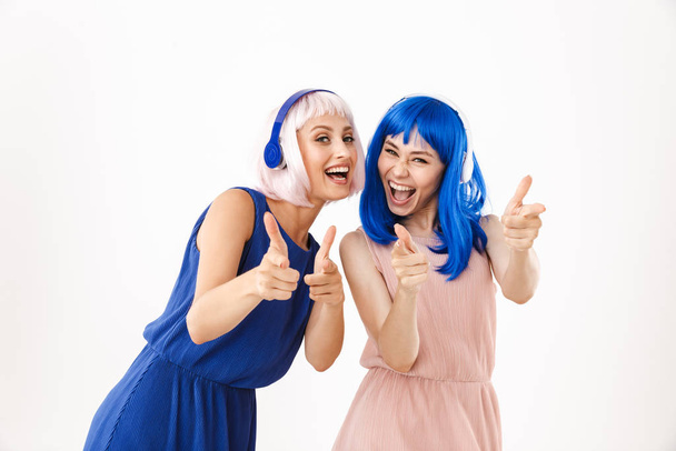 Porträt zweier fröhlicher Frauen mit blauen und rosa Perücken, die mit dem Finger auf die Kamera zeigen, während sie Kopfhörer benutzen - Foto, Bild