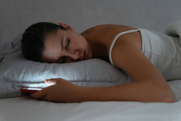 Young woman sleeping on bed with smartphone in her hand late at  - Foto, imagen