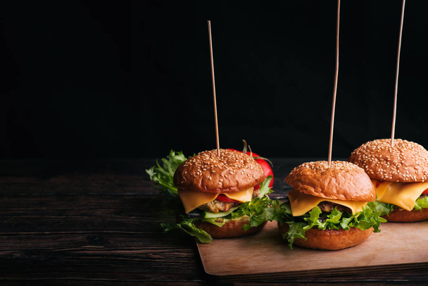 drei hausgemachte Hamburger mit Fleisch, Käse, Salat, Tomate auf einem Holzbrett auf einem Tisch vor schwarzem Hintergrund - Foto, Bild