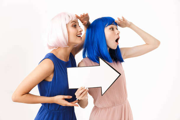 Portrait of two surprised women wearing blue and pink wigs holding blank copyspace arrow pointer - Photo, image