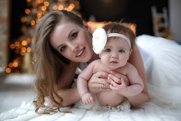 Home portrait of mother and baby on the night before Christmas.Christmas and Happy New Year Holidays. Family, mother and child concept. Happy mom and her cute baby girl in white having fun  sitting together near Christmas tree indoors. - Foto, Bild