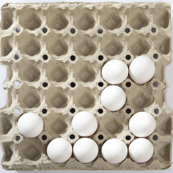 Chicken eggs in a white cardboard container lie on beige wooden kitchen table. - Фото, изображение