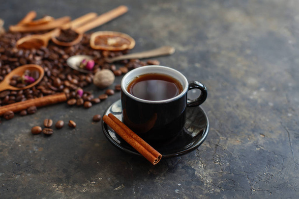 Egy csésze aromás fekete kávé fahéjas bottal a csészealjon. Hozzávalók a kávé készítéséhez a háttérben. Sötét textúrájú kreatív felület. Közelkép, szelektív fókusz. - Fotó, kép