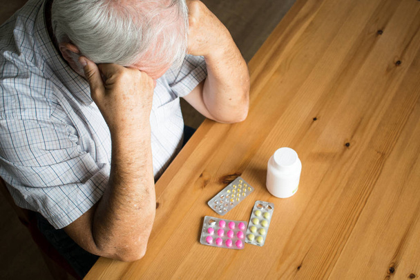 malade homme âgé assis à côté de la table à la maison regarder pilules triste seul
 - Photo, image