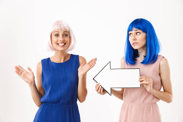 Portrait of two upset and happy women wearing blue and pink wigs holding blank copyspace arrow pointer - Valokuva, kuva