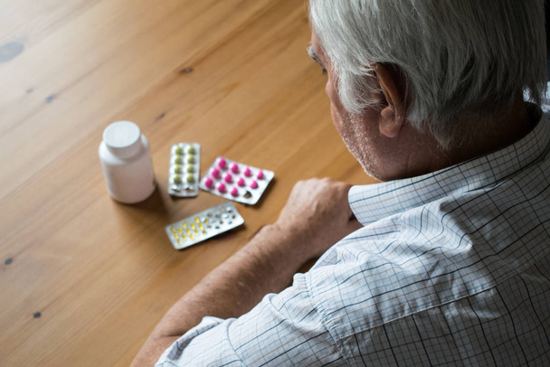 sick senior man sit next to table at home look at pills sad alone - Foto, immagini