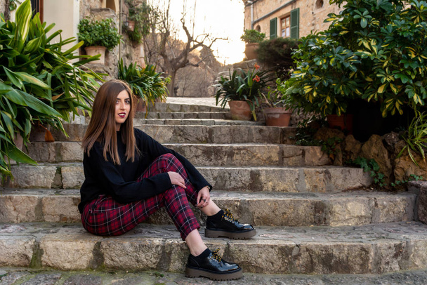 Linda chica de cabello castaño con suéter negro y pantalones rojos sentados en las escaleras en un callejón en el pueblo mallorquín de Valldemossa
 - Foto, imagen