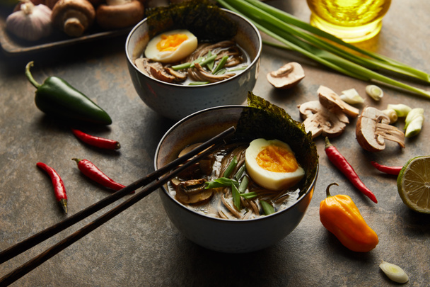 traditional spicy ramen in bowls with chopsticks and vegetables on stone surface - Foto, afbeelding