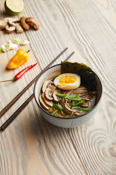 selective focus of traditional spicy ramen in bowl near chopsticks and ingredients on wooden table - Fotografie, Obrázek