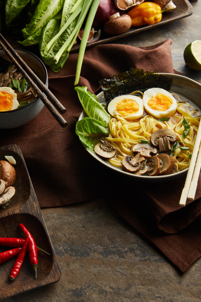 traditional spicy ramen in bowl with chopsticks and ingredients on brown napkin on stone surface - Fotografie, Obrázek