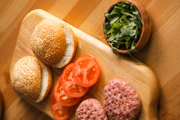 ground beef patties and burger buns with tomato slices on wooden cutting board, close view - Photo, Image