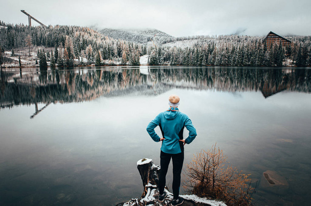 Młody człowiek stojący samotnie i gapiący się na jezioro. Mgła nad wodą. Mgliste powietrze. Wczesny chłodny poranek zimą. Piękny moment wolności i spokojna atmosfera w przyrodzie. Widok z tyłu. - Zdjęcie, obraz