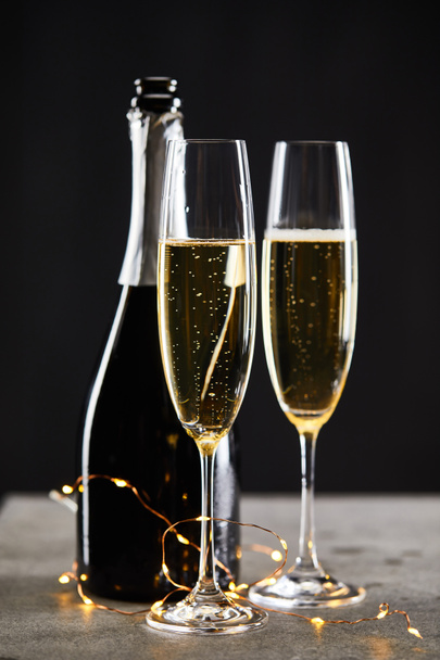 sparkling wine in glasses and bottle with yellow christmas lights, on black - Photo, Image