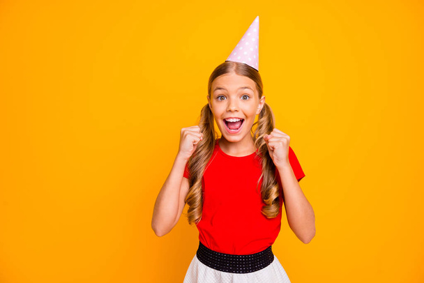 Photo of pretty small lady long tails hairstyle celebrating birthday party good mood raise fists wish open gifts wear paper hat casual red white dress isolated yellow color background - 写真・画像