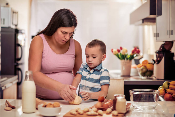 Pregnant mother and her son enjoying in kitchen and making cookies. - 写真・画像